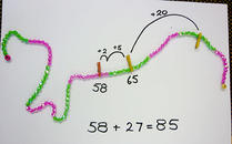 A string of beads, alternating every ten beads from green to pink. The problem is to add 27 to 58. First peg is at 58 with a jump of 2 to reach 60, then a jump of 5 to reach 65 then a jump of 20 to reach the answer of 85.